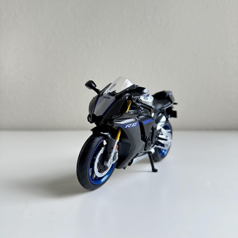 May include: A black and blue Yamaha R1 motorcycle model. The model features a detailed design with blue accents on the wheels and body. The motorcycle has a windshield and a side mirror. The model is on a white surface.