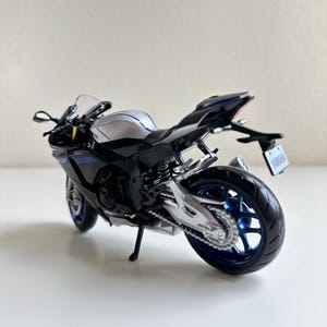 May include: A black and silver Yamaha R1 model motorcycle with blue accents. The model has a license plate that reads "Yamaha". The motorcycle is on a white surface.