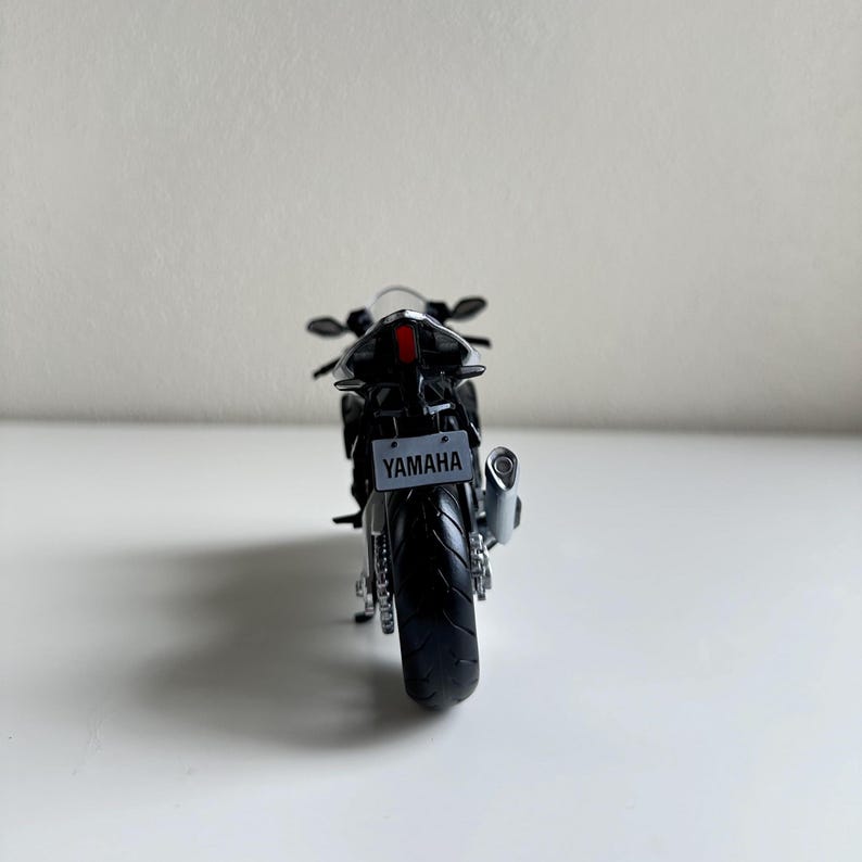 May include: A black Yamaha motorcycle model, viewed from the rear, with a license plate that reads "YAMAHA". The motorcycle has a silver exhaust pipe and a red taillight. The tires are black, and the model is set against a white background.