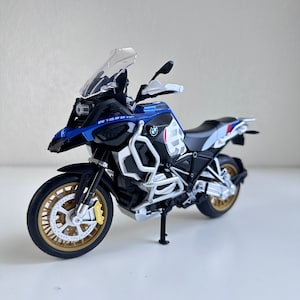 May include: A blue and white BMW R 1250 GS Adventure motorcycle model with a clear windshield. The motorcycle has a black seat and black wheels with gold accents.