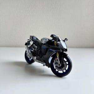 May include: A black and silver Yamaha R1 model motorcycle with blue accents. The model has detailed features including a windshield, mirrors, and a visible engine. The wheels are blue with silver detailing. The motorcycle is on a white surface.