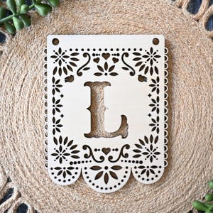 May include: A rectangular light-colored wooden plaque featuring a laser-cut design with a central letter L. The border is decorated with intricate floral patterns, small hearts, and scalloped edges. Two circular holes are positioned at the top corners.