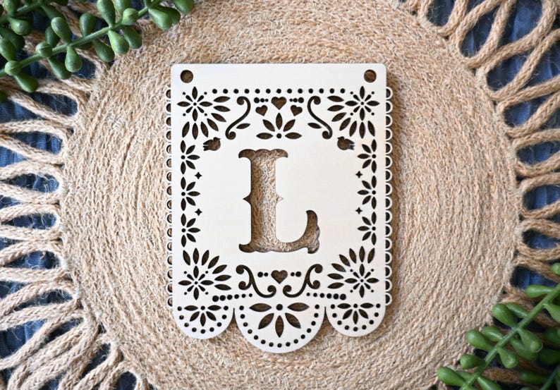 May include: A rectangular light-colored wooden plaque featuring a laser-cut design with a central letter L. The border is decorated with intricate floral patterns, small hearts, and scalloped edges. Two circular holes are positioned at the top corners.