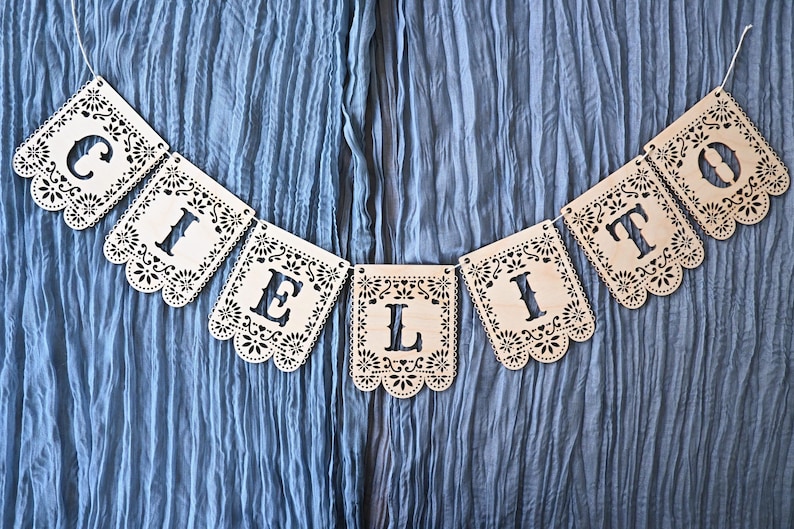 May include: A decorative banner made of eight light-colored wooden panels, each featuring intricate laser-cut floral patterns and scalloped edges. The panels are strung together to spell out the word CIELITO in bold, cut-out capital letters.