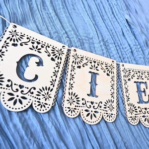 May include: A light-colored wooden banner featuring laser-cut rectangular panels with intricate floral and scalloped edges. Each panel displays a large, bold cutout letter, specifically the letters C, I, and E, strung together on a thin white cord.