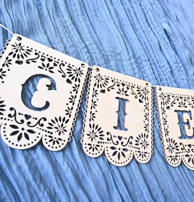 May include: A light-colored wooden banner featuring laser-cut rectangular panels with intricate floral and scalloped edges. Each panel displays a large, bold cutout letter, specifically the letters C, I, and E, strung together on a thin white cord.