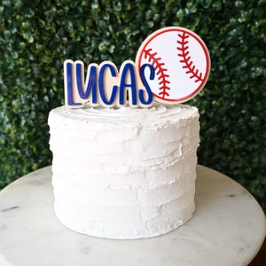 May include: A white frosted cake with a baseball-themed topper. The topper features the name "LUCAS" in blue letters and a baseball design. The cake sits on a marble cake stand against a green hedge background.