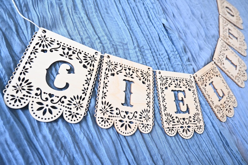 May include: A decorative banner made of light-colored wood panels, each featuring intricate laser-cut floral patterns and scalloped edges. The panels are strung together to spell out the word CIELITO in bold, cut-out capital letters.