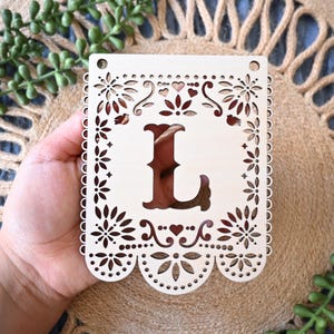May include: A rectangular, light-colored wooden plaque featuring a laser-cut design with a large central letter L. The border is decorated with intricate floral patterns, small hearts, and scalloped edges at the bottom, with two holes at the top.