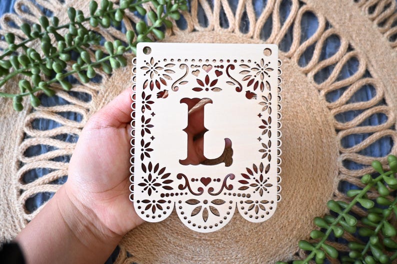 May include: A rectangular, light-colored wooden plaque featuring a laser-cut design with a large central letter L. The border is decorated with intricate floral patterns, small hearts, and scalloped edges at the bottom, with two holes at the top.