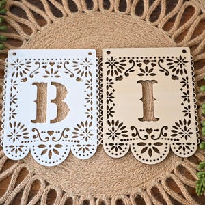 May include: Two rectangular laser-cut wooden banner flags featuring intricate floral and heart patterns along the edges. One flag is painted white with the letter B, and the other is natural light wood with the letter I. Both have two holes at the top.