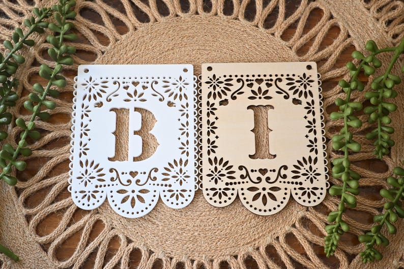 May include: Two rectangular laser-cut wooden banner flags featuring intricate floral and heart patterns along the edges. One flag is painted white with the letter B, and the other is natural light wood with the letter I. Both have two holes at the top.