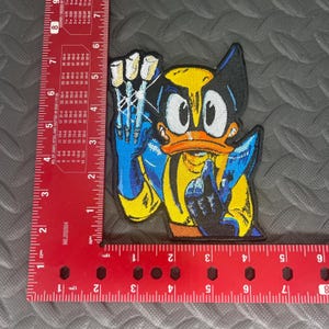 May include: A yellow and blue embroidered patch of a cartoon duck dressed as Wolverine, with three claws extended from each hand. The duck has a yellow beak and large, round eyes.