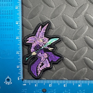 May include: An embroidered patch featuring a purple-clad wizard character holding a glowing green staff. The patch is positioned against a blue L-shaped ruler, showing a height of approximately 3.5 inches and a width of approximately 3 inches.