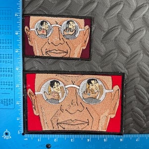 May include: Two embroidered patches featuring a stylized face wearing glasses. The glasses reflect a person in a tan jacket. The patches have a red border and black trim. The larger patch measures approximately 4 inches by 3 inches.