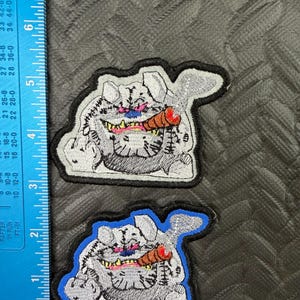 May include: Two embroidered patches featuring a cartoonish, gray creature with a cigar. The creature has a wide grin, pink tongue, and red lips. One patch has a black border, the other a blue border. The patches are on a textured gray surface.