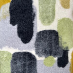 May include: Close-up of a soft, textured fabric with an abstract pattern. The design features irregular shapes in shades of gray, green, and yellow against a cream-colored background. The fabric has a plush, fuzzy appearance, suggesting a cozy textile.