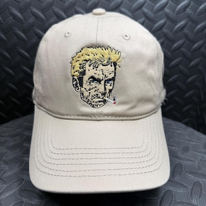 May include: A beige baseball cap featuring an embroidered graphic of a man with blonde hair and a cigarette in his mouth. The design uses black, yellow, white, and red thread to detail the facial features and hair on the front of the cap.