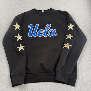 May include: Black crewneck sweatshirt with the word "Ucla" in blue script with a white outline. Gold glitter star patches are on both sleeves. The sweatshirt is a casual style.