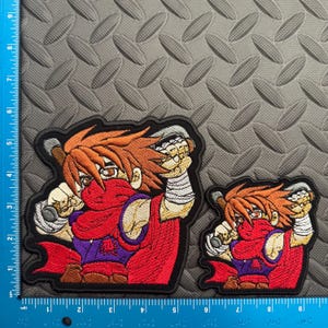 May include: Two embroidered patches featuring a cartoon character in a fighting stance. The character has orange hair, a red scarf, and a purple top. The patches are outlined in black and are approximately 4 inches and 2 inches in height.