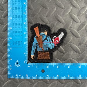 May include: An embroidered patch featuring a character with a brown shotgun strapped to their back and a gray chainsaw attached to their right arm. The character wears a blue shirt and brown pants. A blue L-shaped ruler shows the patch is 3 inches tall.