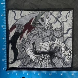 May include: Embroidered square patch featuring a detailed illustration of a warrior with a horned helmet, wings, and clawed hands. The design uses shades of gray, black, and burgundy. The patch is approximately 4 inches square.