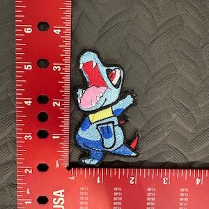 May include: A blue and red embroidered patch of a cartoon dinosaur with a yellow collar. The dinosaur is roaring with its mouth open and has a pink tongue.