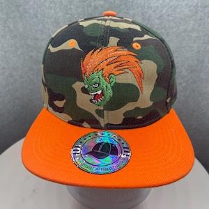 May include: Camouflage print snapback hat with an orange brim and top button. The hat features an embroidered green and orange character design. A holographic sticker with the words "Original Snapbacks" is on the brim.