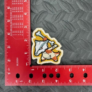 May include: An embroidered iron-on patch featuring a cartoon dog wearing a hat and holding a bone. The dog is brown and white, and the hat is gray. The background of the patch is yellow.