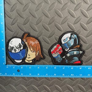 May include: Two embroidered patches placed next to a blue ruler. The left patch shows a person with brown hair holding a skull mask with a blue chin area. The right patch depicts a character in a black and blue armored suit with a red glove.