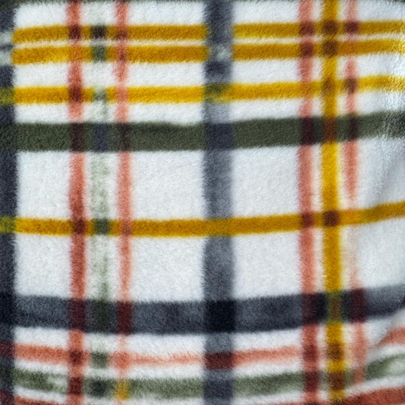 May include: Close-up of a soft, white fleece fabric with a plaid pattern. The plaid design features intersecting lines in shades of yellow, orange, green, gray, and brown, creating a textured, cozy appearance. The fabric is likely used for blankets or throws.