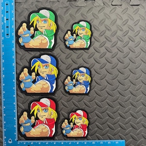 May include: Embroidered patches featuring a cartoon character in various color schemes: green and white, blue and white, and red and white. The character has yellow hair, blue eyes, and is pointing forward. Patches are in two sizes.