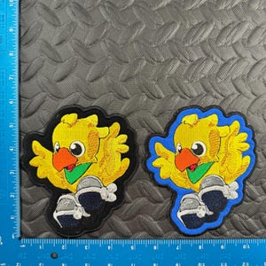 May include: Two embroidered patches featuring a yellow cartoon bird with an orange beak, green scarf, and blue and silver boots. One patch has a black border, the other a blue border. The patches are approximately 3 inches wide.