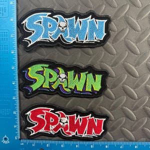 May include: Three embroidered patches with the word "SPAWN" in blue, green, and red, each with a skull graphic. The patches are outlined in black and white.