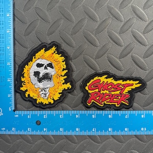 May include: Two embroidered patches featuring a skull surrounded by yellow and orange flames and the text Ghost Rider in red with yellow flames. A blue L-shaped ruler shows the skull patch is 3 inches tall and the text patch is 4 inches wide.