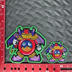 May include: Two embroidered patches featuring a cartoon character. The larger patch is green, purple, red, yellow, and blue. The smaller patch is the same design but smaller. Both patches feature a character that looks like a clock with wings and a crown.