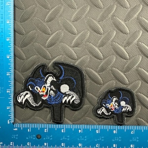 May include: Two embroidered patches featuring a cartoon demon with black wings, blue accents, and white claws. The larger patch measures approximately 3 inches wide. The patches are on a textured gray surface with a ruler for scale.