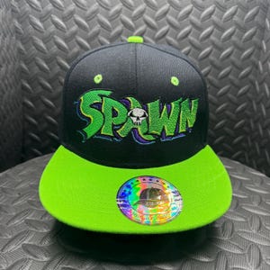 May include: Black and neon green Spawn baseball cap. The cap features the word "SPAWN" in neon green with a skull graphic. The brim is neon green, and the cap has a round sticker with a hat graphic.