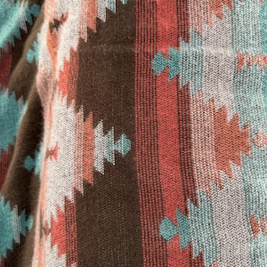 May include: Close-up of a woven textile with a geometric pattern. The design features repeating shapes in shades of brown, teal, red, and off-white. The fabric has a textured appearance, suggesting a soft, warm material. The overall aesthetic is reminiscent of Southwestern or tribal designs.