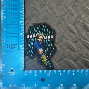 May include: Embroidered patch featuring a man with the text "BABA YAGA" in white. The man is depicted in a blue jacket, holding a gun, with a rain and lightning design. The patch has a black border and is on a gray surface.