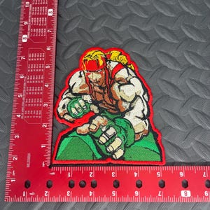 May include: Embroidered patch featuring a red-headed, muscular character with a red headband, green and white clothing, and a red border. The character is in a fighting pose with a green and white glove.