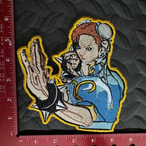 May include: Embroidered patch featuring a character from a video game. The character is a woman with brown hair, wearing a blue and white outfit with a yellow trim. She is in a fighting pose with her right arm extended and her left hand clenched in a fist.