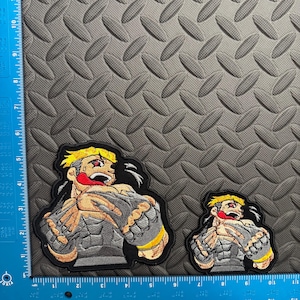 May include: Two embroidered patches featuring a muscular cartoon character with yellow hair, red eyes, and a red tongue. The character is wearing gray and yellow armor. The patches are on a gray textured surface, with a ruler in inches.