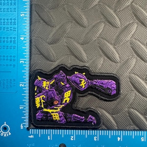 May include: Embroidered patch featuring a purple robot design with yellow accents and Japanese text. The patch is outlined in black and measures approximately 2.5 inches wide and 2 inches tall.