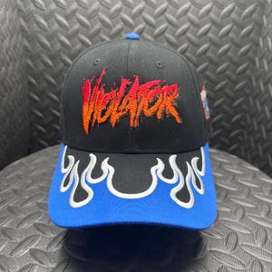 May include: Black baseball cap with a blue brim and a flame design. The word "VIOLATOR" is embroidered in orange and red on the front. The cap has a blue button on top.