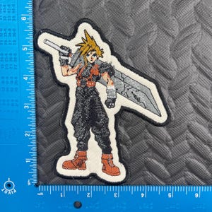 May include: Embroidered patch featuring a character with spiky blonde hair, wearing a black outfit, and holding a large sword. The patch has a white border and is on a gray surface. The character is in a fighting stance.