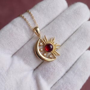 May include: A gold pendant necklace featuring a crescent moon and sun motif. The center of the sun is a round, deep red gemstone. Three small, clear, round-cut stones are set along the curve of the moon. The pendant hangs from a delicate gold chain.