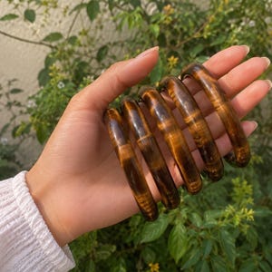 Puede incluir: Un conjunto de cinco brazaletes de piedra ojo de tigre, pulidos y lisos, que presentan un caracter&iacute;stico patr&oacute;n de bandas de color marr&oacute;n dorado chatoyante. Cada pulsera circular muestra un acabado brillante con variaciones naturales.