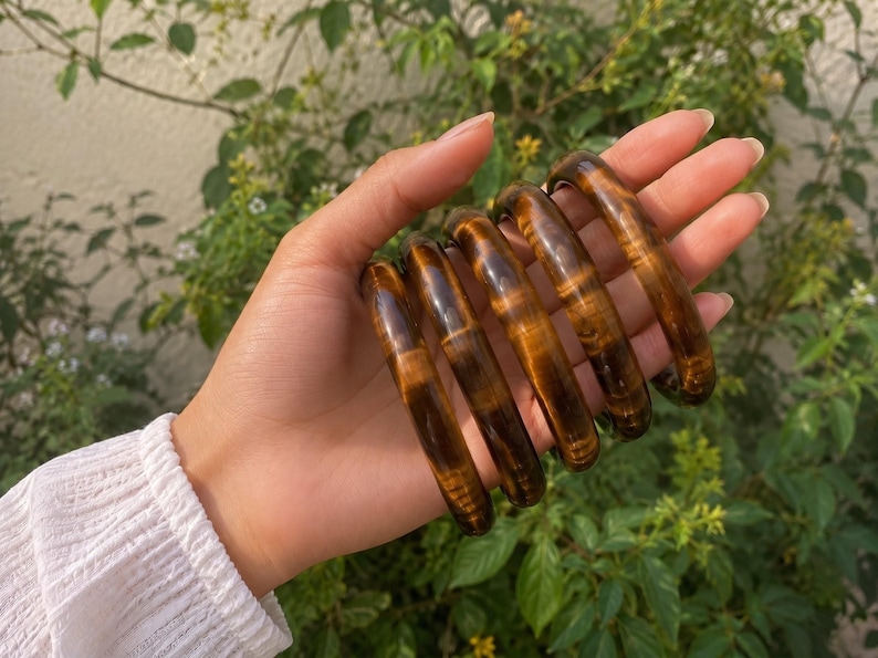 Puede incluir: Un conjunto de cinco brazaletes de piedra ojo de tigre, pulidos y lisos, que presentan un caracter&iacute;stico patr&oacute;n de bandas de color marr&oacute;n dorado chatoyante. Cada pulsera circular muestra un acabado brillante con variaciones naturales.