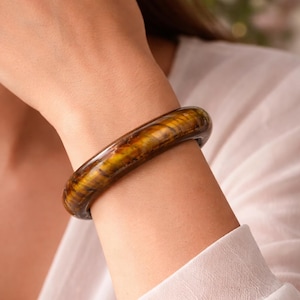 Puede incluir: Un brazalete r&iacute;gido pulido hecho de piedra ojo de tigre, con una superficie lisa y redondeada que presenta bandas chatoyantes caracter&iacute;sticas en tonos marr&oacute;n dorado y marr&oacute;n oscuro, creando un aspecto brillante y estratificado en la mu&ntilde;eca.